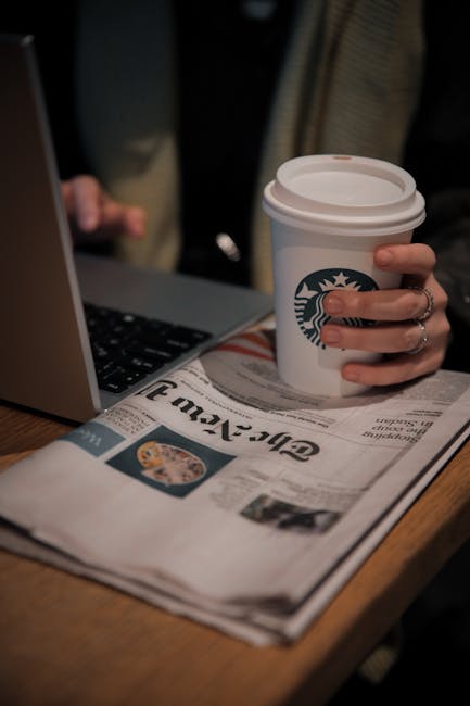 Persona che lavora al computer con caffè e giornale, esempio di integrazione tecnologia e informazione nella vita quotidiana.