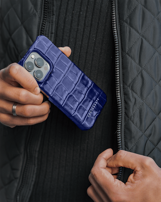 Custodia per iPhone in pelle blu effetto coccodrillo tenuta in mano da un uomo, dettaglio elegante e protezione per smartphone