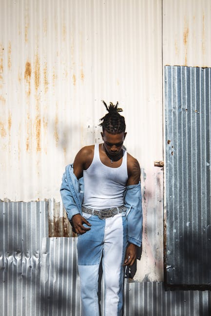 Uomo con dreadlocks e abbigliamento denim appoggiato a parete metallica, stile urbano e moderno, espressione pensierosa.