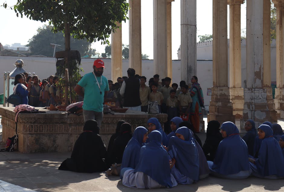 Studenti in abiti religiosi blu seduti a terra, con un uomo in maglietta verde e cappellino rosso in piedi, in un contesto scolastico.