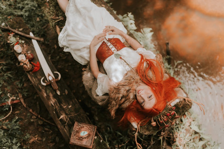 Giovane donna con capelli rossi, vestito bianco e corpetto sdraiata su un tronco con spada e libro magico, evocando l'epica greca.