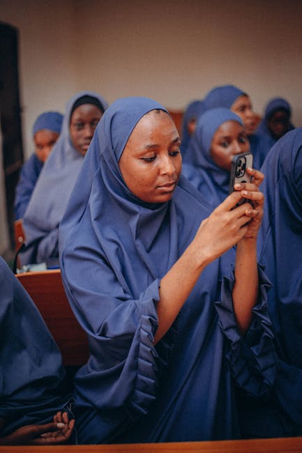 Studentessa con hijab usa smartphone in classe, riflettendo il dibattito sulla digitalizzazione scolastica e l'uso dei cellulari.