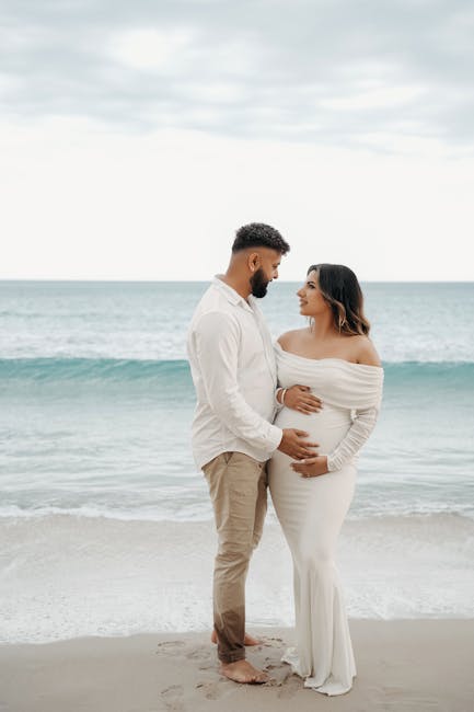 Coppia in attesa sulla spiaggia con abito bianco: celebrazione della gravidanza e dell'amore in riva al mare, futuro genitoriale.