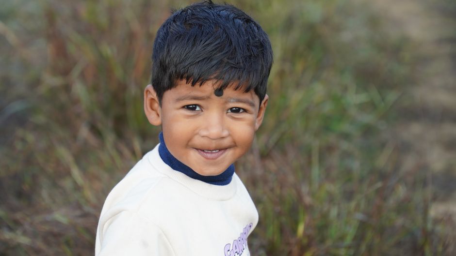 Ritratto di un bambino sorridente con un punto nero sulla fronte, vestito con una maglietta bianca e collo blu, sullo sfondo di erba secca.