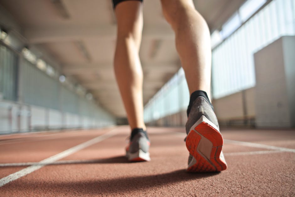 Gambe di un corridore su pista rossa, scarpe da ginnastica grigie e arancioni, vista dal basso. Metafora di velocità e progresso.
