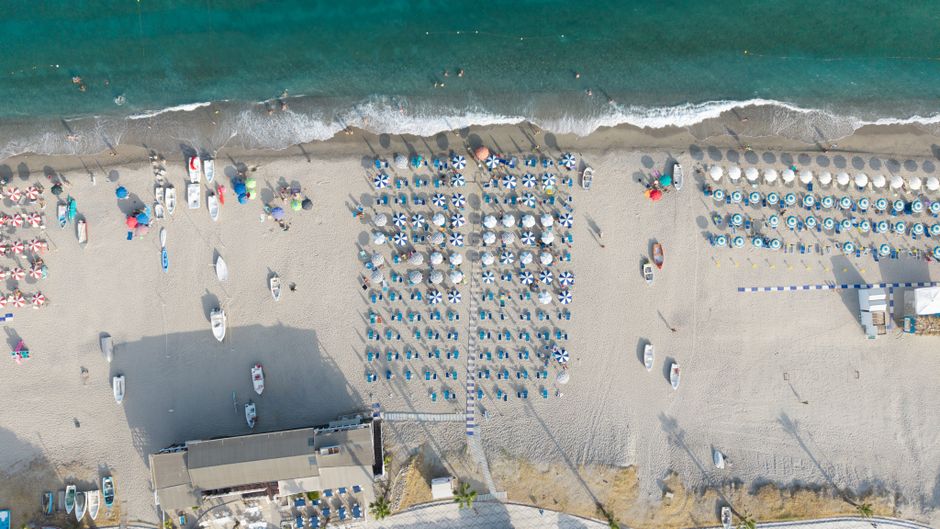 Vista aerea di una spiaggia affollata con ombrelloni e lettini, con il mare turchese sullo sfondo, in riferimento al caldo a Catanzaro e alla chiusura anticipata delle scuole.