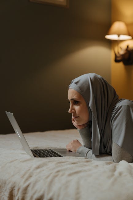 Donna musulmana con hijab grigio che usa un laptop a letto, riflessione sulla proposta di legge contro il separatismo islamico.