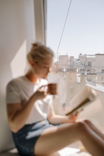 Studentessa bionda legge un libro e beve caffè vicino alla finestra con vista sulla città, profilo dello studente e relax
