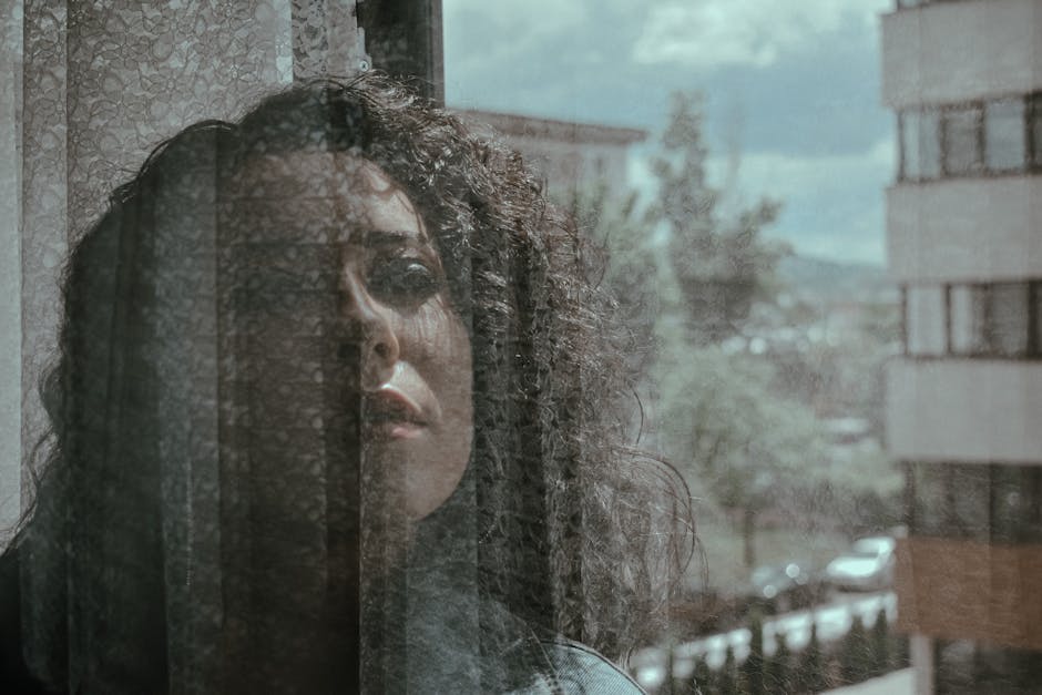 Ritratto di donna pensierosa dietro una tenda, riflesso della città sullo sfondo, evocando la perdita e la malinconia urbana italiana.