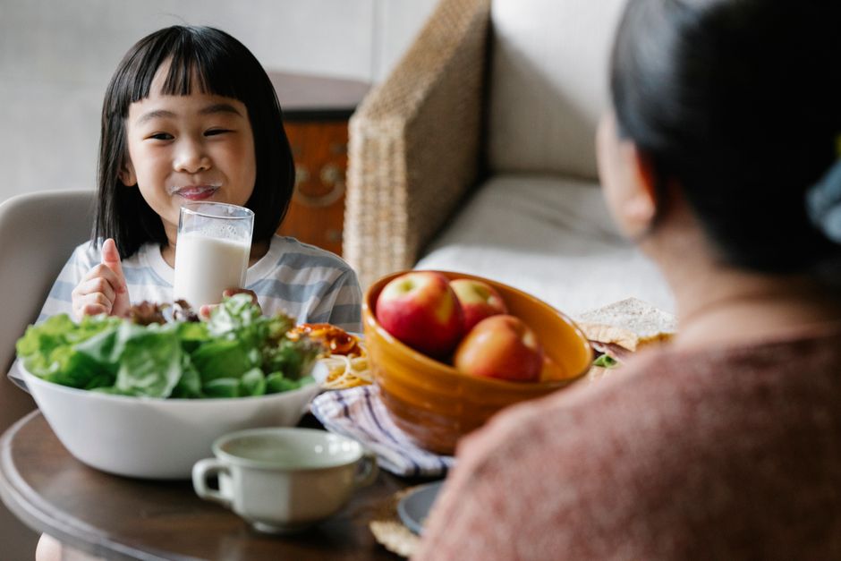 Bambina sorridente beve latte da un bicchiere, con un pasto sano a base di insalata e frutta, promuovendo abitudini alimentari corrette nelle scuole.