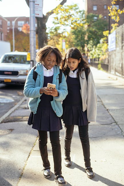 Due studentesse in uniforme guardano un cellulare per strada, riflettendo sul divieto di utilizzo a scuola e l'importanza dell'educazione digitale.