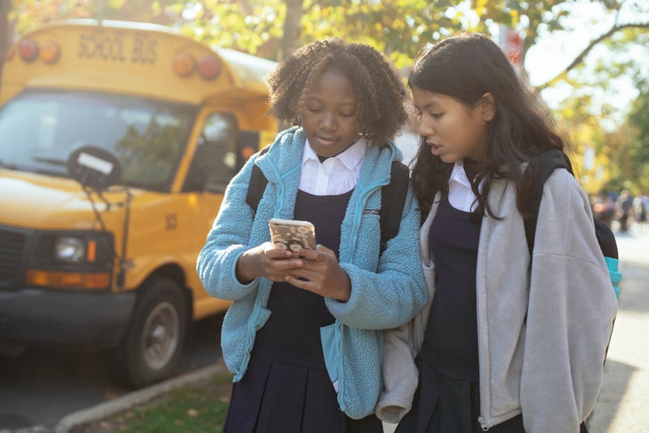 Due studentesse controllano lo smartphone davanti allo scuolabus giallo, guida alla domanda di iscrizione scolastica efficace.