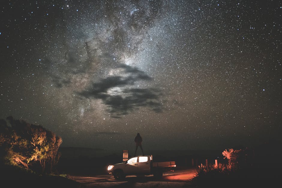 Uomo in piedi su un pick-up sotto un cielo stellato mozzafiato, esplorando il cosmo e la Via Lattea in una notte buia e suggestiva.