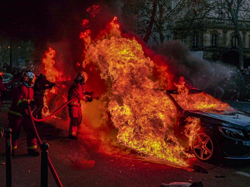 Incendio auto durante proteste: immagine di violenza urbana con vigili del fuoco al lavoro per spegnere le fiamme in strada.