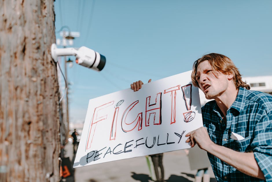 Manifestante con cartello 'Fight Peacefully' sotto telecamera, simbolo di lotta pacifica per la giustizia sociale e sorveglianza