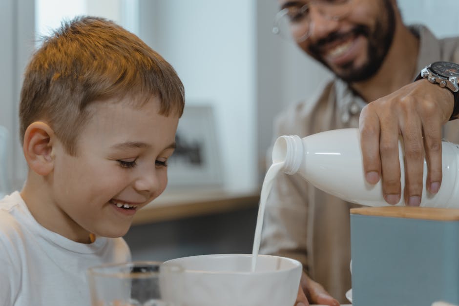 Bambino sorridente guarda il latte versato nella ciotola, un momento di gioia e felicità condiviso con il padre durante la colazione