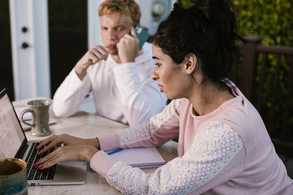 Insegnante concentrata al laptop mentre collega ascolta al telefono, ottimizzazione tempo e risorse per docenti impegnati