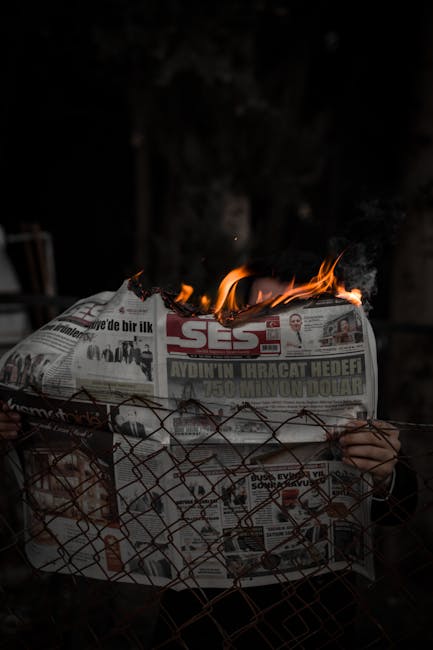 Giornale in fiamme dietro una recinzione metallica, simbolo di disinformazione e crisi mediatica legata alla vicenda di Crans-Montana.