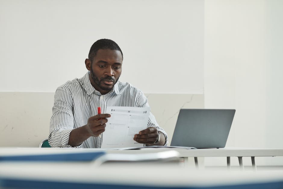 Insegnante afroamericano esamina documenti con penna rossa e laptop, valutazione didattica per insegnamento efficace