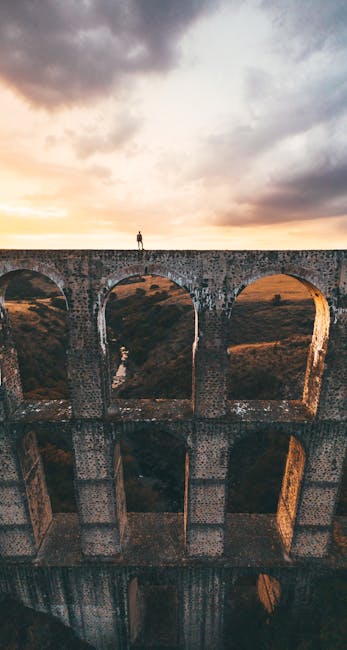 Uomo in piedi sull'imponente diga ad archi, simbolo di ingegneria e potenza, con cielo al tramonto che esalta la sua maestosità.