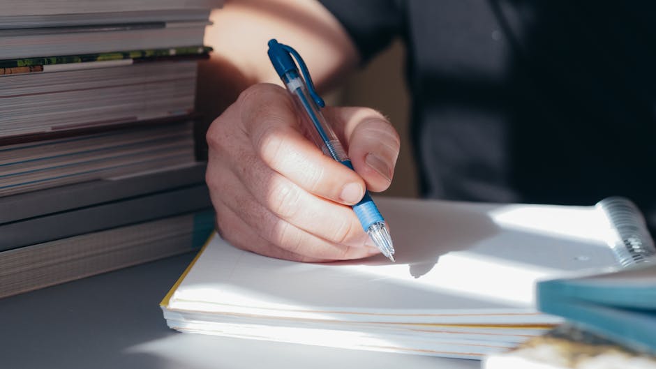 Studente scrive su quaderno con penna blu, libri impilati sullo sfondo. Investimenti Umbria per il diritto allo studio e laboratori di scrittura.