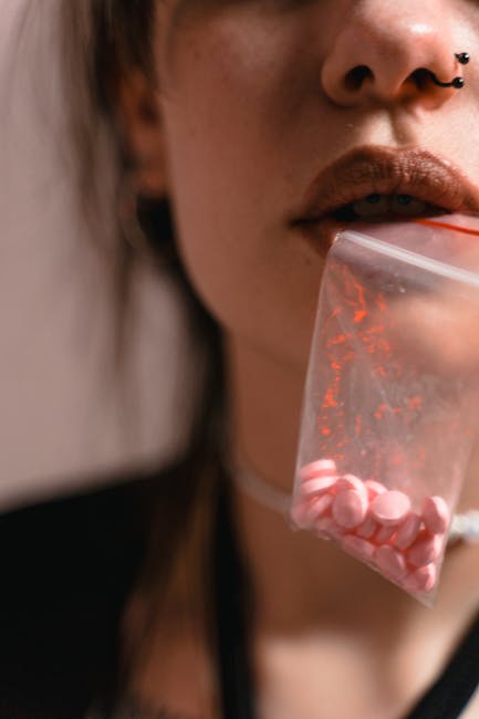 Giovane donna con piercing al naso tiene in bocca una bustina di pillole rosa, evocando riflessioni sulla bellezza e la decadenza nella cultura contemporanea.