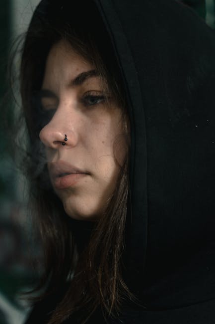 Giovane donna pensierosa con piercing al naso e felpa con cappuccio nera, simbolo di introspezione e scoperta del proprio cammino personale e valori unici.