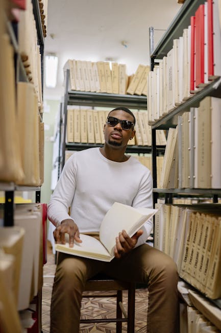 Uomo con occhiali da sole consulta archivio tracce esami di stato seduto tra scaffali pieni di documenti e fascicoli