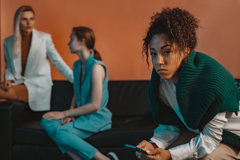 Incertezza e relazioni: donna pensierosa con smartphone, sullo sfondo altre due donne sedute su un divano. Riflessioni sulla fiducia.