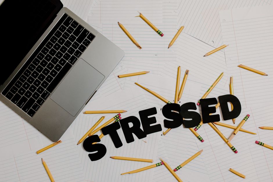 Lettere nere formano la parola STRESSED su fogli a righe, circondate da matite spezzate e un laptop, simbolo di stress lavorativo per docenti e ATA.