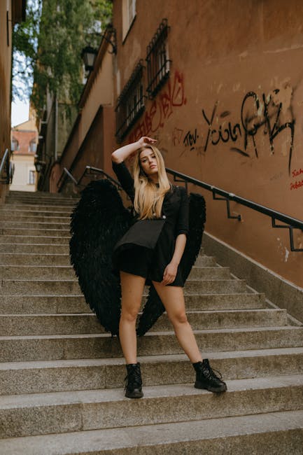 Donna bionda con ali nere in posa su scalinata urbana con graffiti, look dark angel e stivali anfibi neri, atmosfera misteriosa