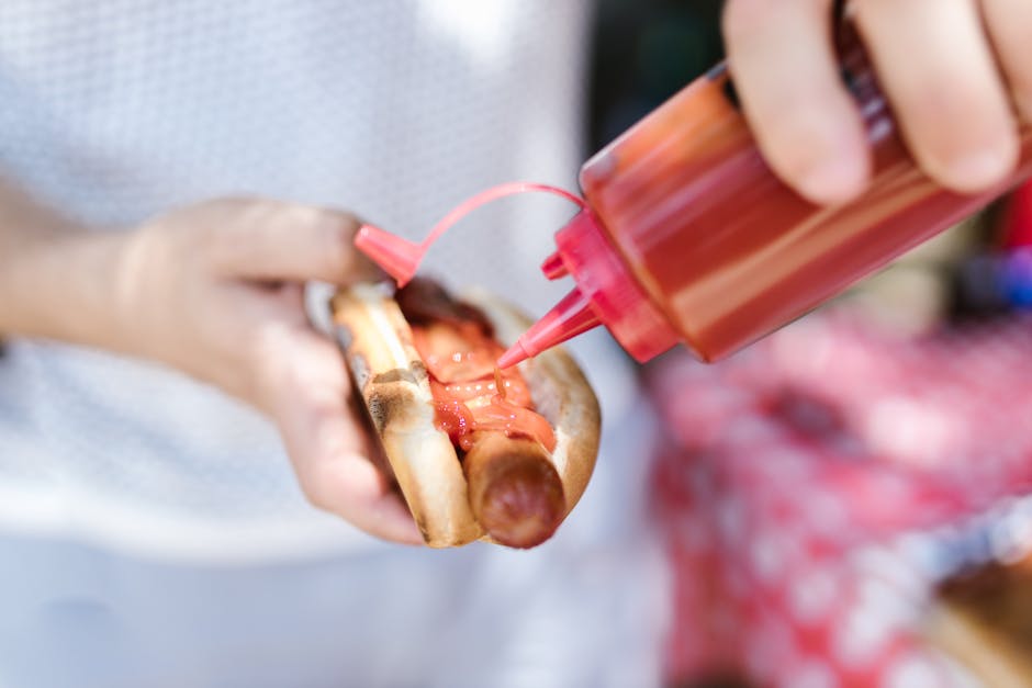 Hot dog con ketchup versato da flacone rosso, mani che lo tengono, focus sul condimento e sul panino