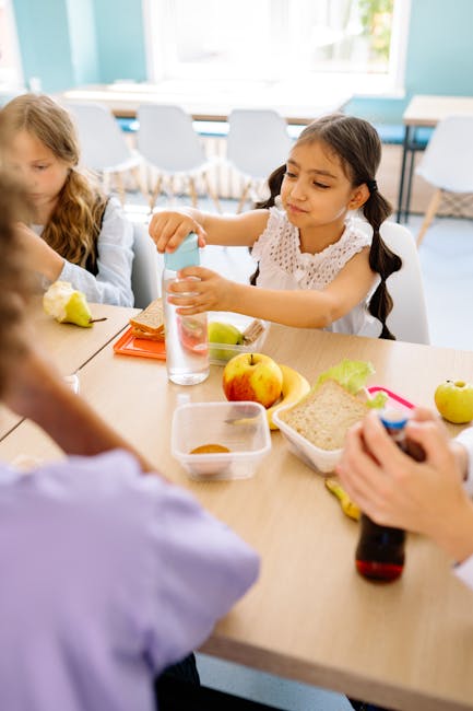 Bambina a mensa apre la borraccia con cibo scolastico intorno, evocando ricordi di infanzia legati alla mensa e alla maestra.