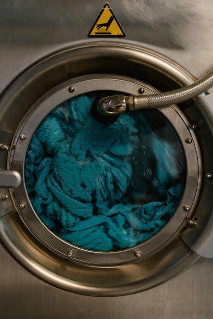 Ciclo operativo lavatrice industriale: biancheria turchese in movimento durante la fase di lavaggio, dettaglio oblò e avviso di sicurezza.