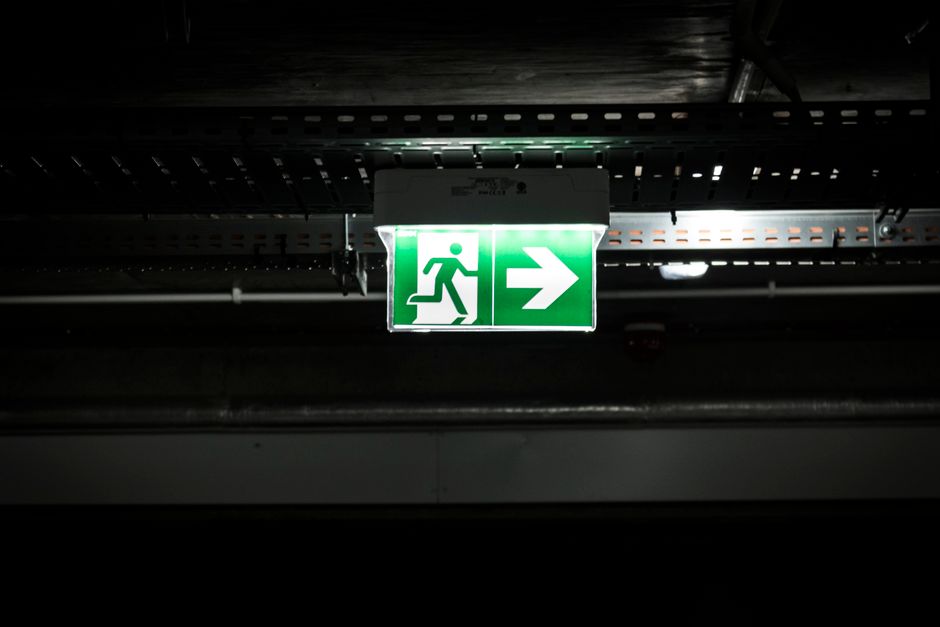 Segnale di uscita di emergenza verde acceso con figura stilizzata che corre verso una freccia, installato in un ambiente industriale scuro, simboleggia la sicurezza e l'evacuazione in caso di emergenza