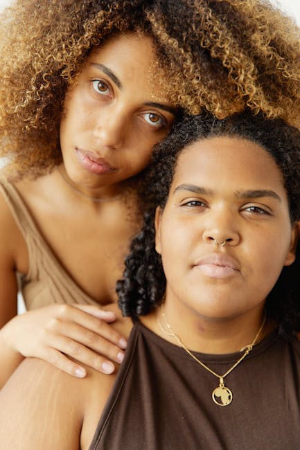 Due giovani donne afroamericane, una con capelli ricci biondi, l'altra con collana a forma di mondo, riflettono fiducia e connessione nell'era digitale.