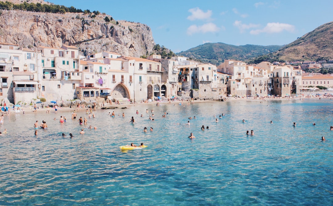Persone che nuotano e si rilassano in mare davanti a un borgo siciliano con case bianche e montagne sullo sfondo, in una giornata di sole