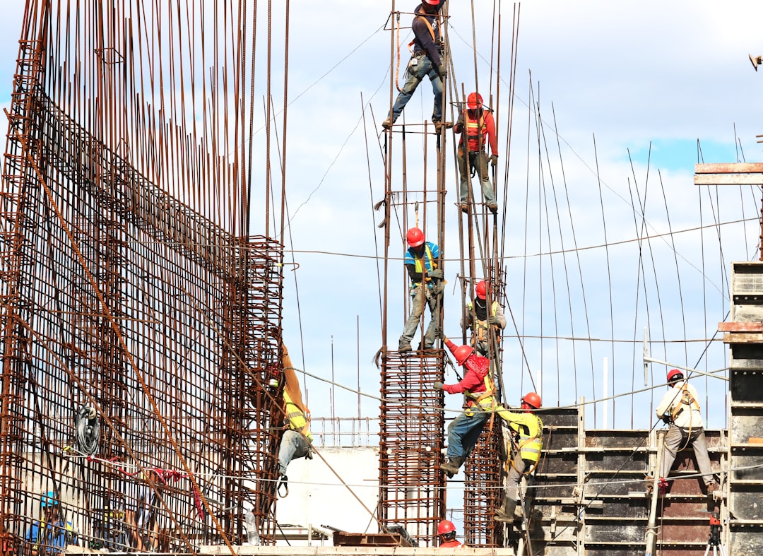 Operai al lavoro su un'impalcatura di un cantiere edile per la costruzione di una scuola, con armature metalliche in primo piano e cielo azzurro sullo sfondo