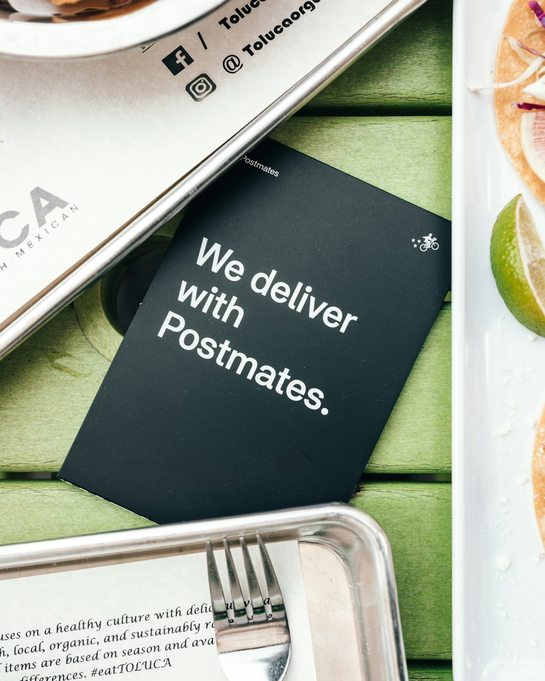 Biglietto nero con scritta We deliver with Postmates, simbolo di consegna cibo e logo Postmates, su un tavolo verde con vassoi metallici e cibo