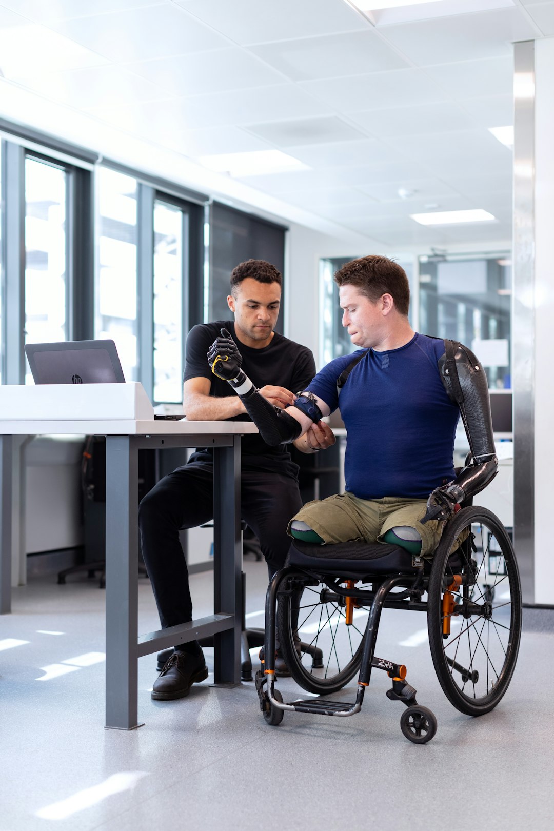 Uomo in carrozzina con protesi bionica al braccio viene assistito da un tecnico durante un corso MIM per la Riforma Disabilità 2027.