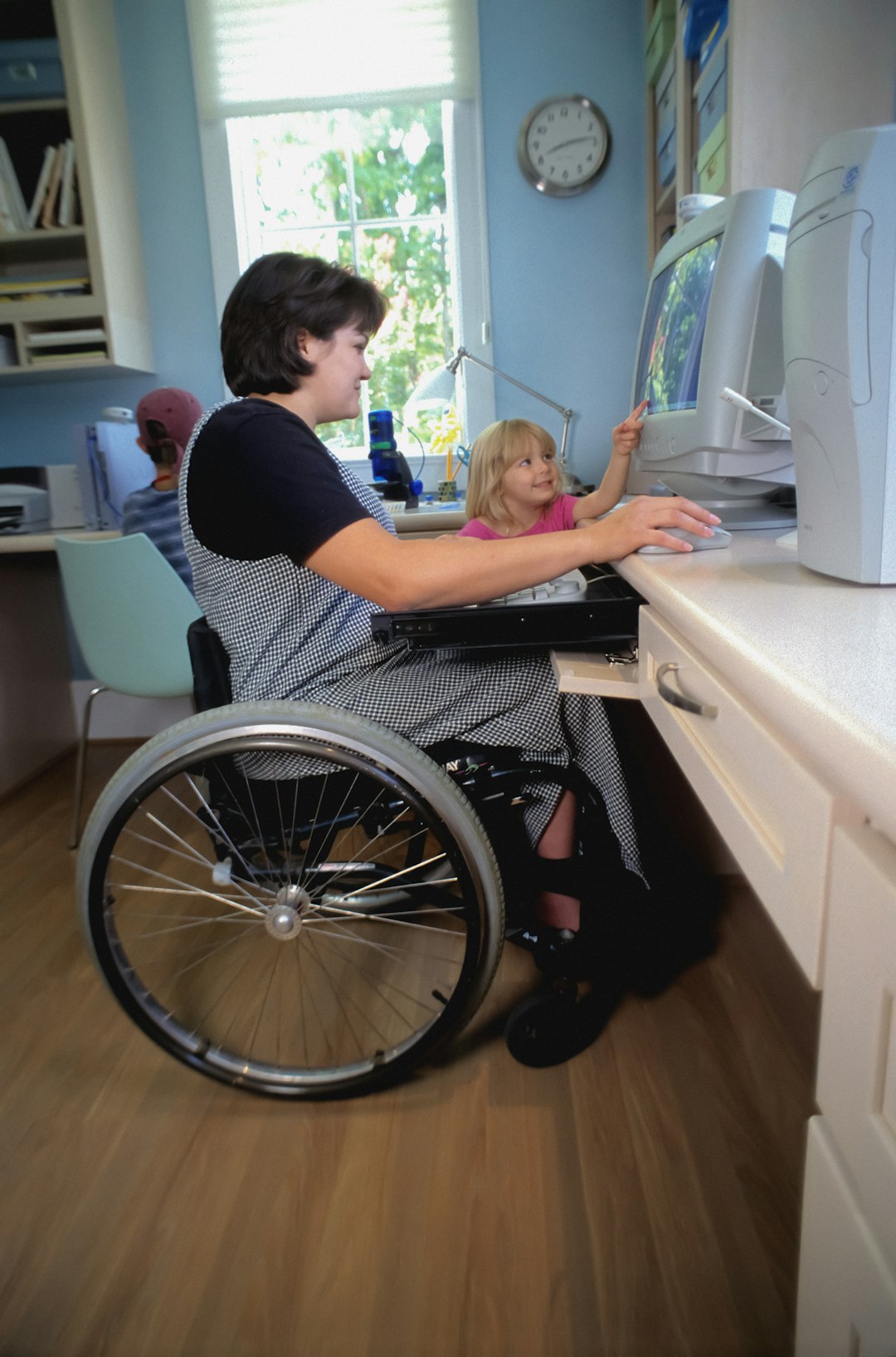 Bambina indica schermo computer mentre donna in sedia a rotelle lavora, supporto apprendimento e inclusione per bambini con difficoltà.