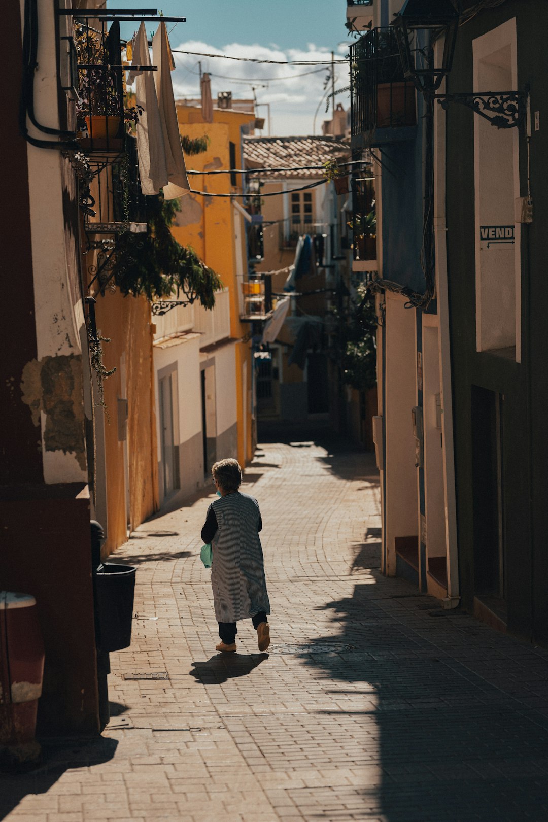 Anziana che cammina sola in una via stretta e assolata di un paese europeo, simbolo di solitudine e rischio povertà per gli anziani.