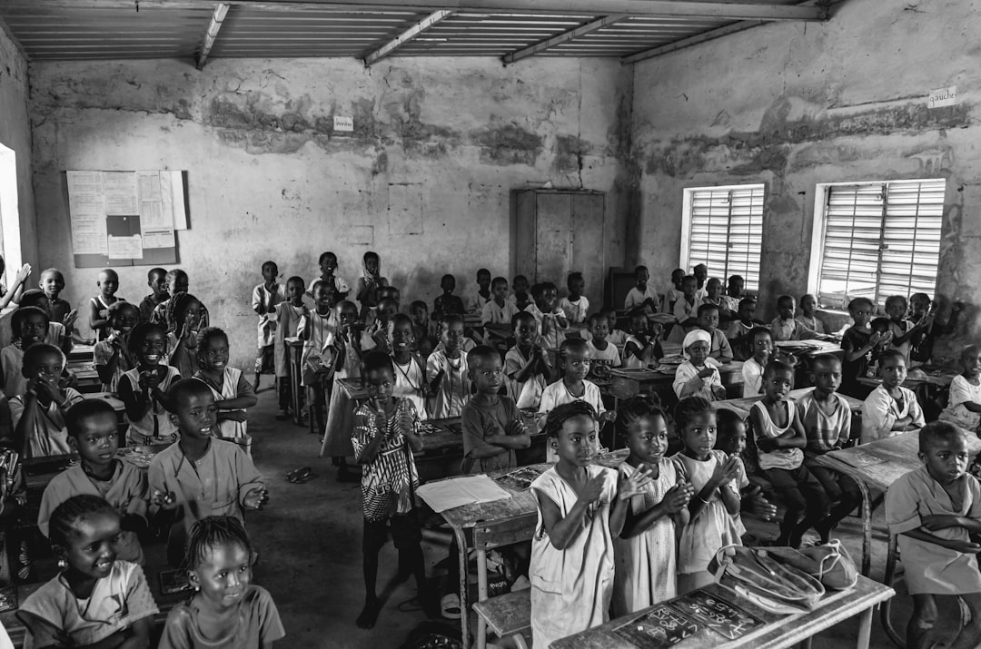 Bambini in aula scolastica con mani alzate, alcuni applaudono, in un ambiente semplice con banchi e lavagne