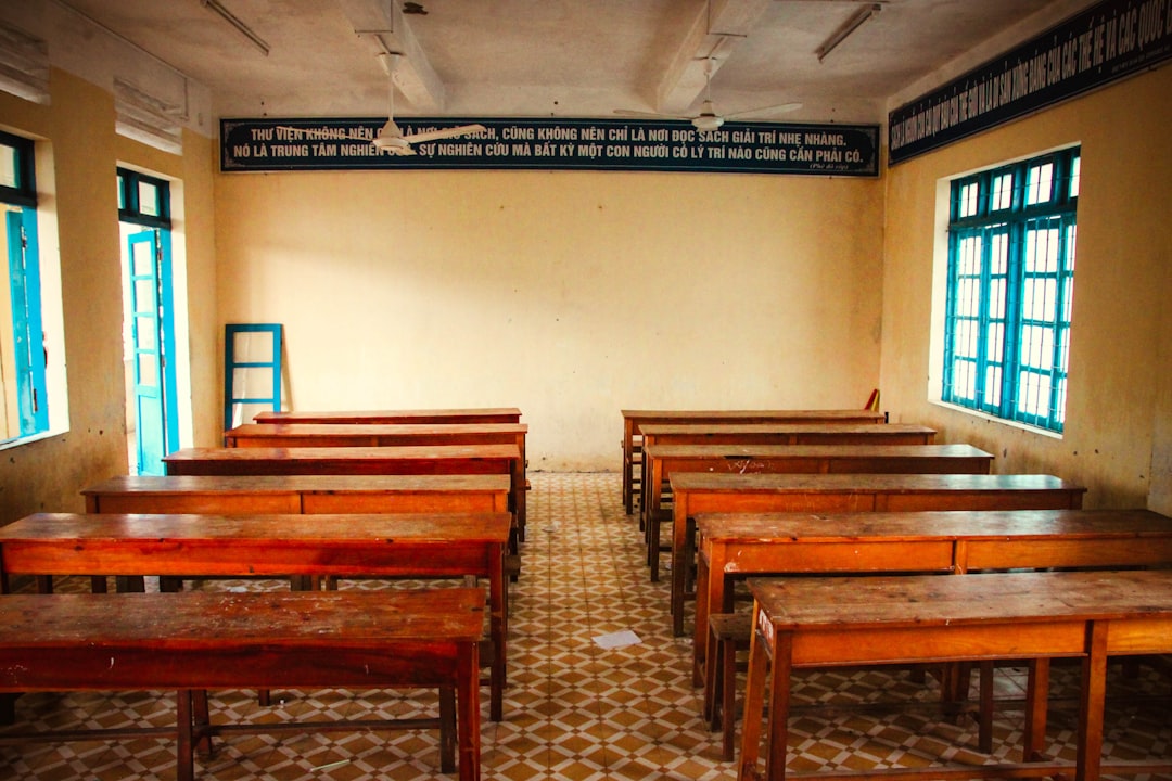 Aula scolastica vuota con banchi in legno e finestre azzurre. Immagine evocativa delle difficoltà nelle visite scolastiche.