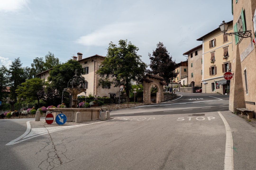 Fontana e arco in pietra in un paese trentino, con segnali stradali che indicano stop e divieto di accesso, simbolo di rinvio delle riforme formative.